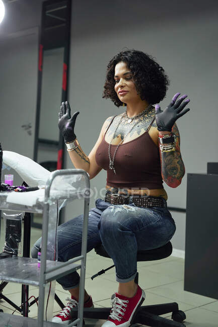 Adult woman in casual clothes with tattoos sitting in light tattoo salon while looking down — Stock Photo