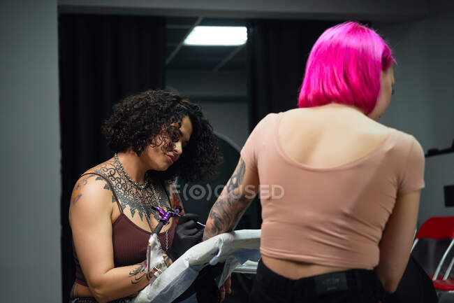 Maestro del tatuaje en guantes usando la máquina profesional del tatuaje mientras que pinta el tatuaje en el brazo de la mujer en el estudio moderno del tatuaje - foto de stock