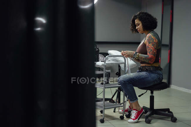 Mulher adulta séria em roupas casuais com tatuagens sentadas no salão de tatuagem leve enquanto olha para baixo — Fotografia de Stock