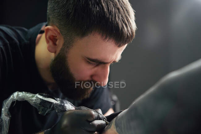 Концентрированный мужчина-татуировщик в перчатках делает татуировку на руке клиента при использовании профессиональной тату-машины в современной тату-студии — стоковое фото
