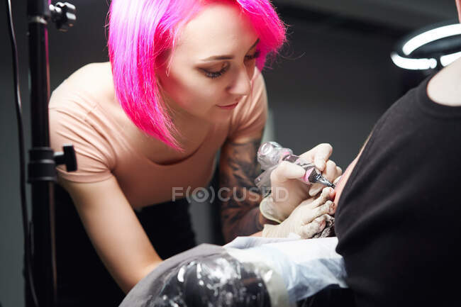 Майстер татуювання з рожевим волоссям в рукавичках, використовуючи професійну татуювальну машину, роблячи татуювання на плечі клієнта в сучасному татуювальному салоні — стокове фото