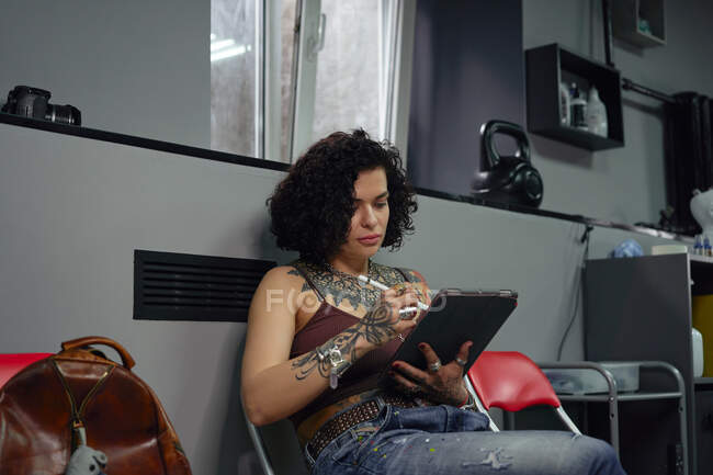 Концентрований майстер татуювання в повсякденному одязі, сидячи на стільці з планшетом і ручкою під час малювання ескізів татуювання в легкому татуюванні салоні — стокове фото