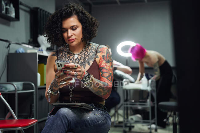 Mulher adulta confiante em roupa casual com tatuagens sentadas com tablet enquanto navega no smartphone no salão de tatuagem brilhante perto de tatuagem fazendo tatuagem para o cliente — Fotografia de Stock