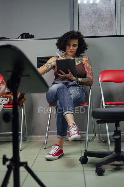 Konzentrierter Tätowiermeister in lässiger Kleidung sitzt mit Tablet und Stift auf einem Stuhl und zeichnet Tätowierskizzen im hellen Tätowiersalon — Stockfoto