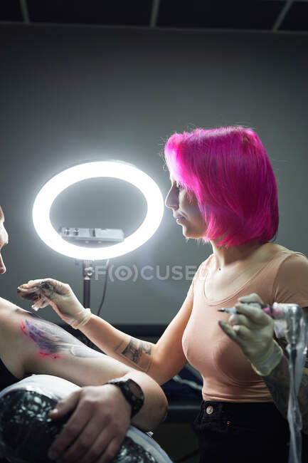 Serious tattoo master in gloves with tattoo machine disinfecting client shoulder before making tattoo in light tattoo studio near ring lamp — Stock Photo
