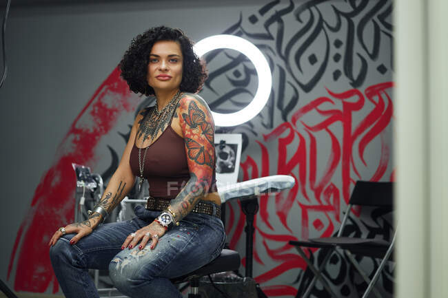 Serious adult woman in casual clothes with tattoos sitting in light tattoo salon while looking at camera — Stock Photo