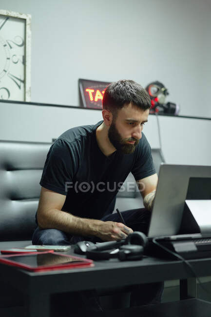 Thoughtful adult guy in casual outfit with tattoos sketching on computer in bright tattoo studio — Stock Photo
