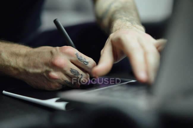 Crop guy en tenue décontractée avec des tatouages esquissant sur ordinateur dans un studio de tatouage lumineux près du tatoueur faisant le tatouage pour le client — Photo de stock