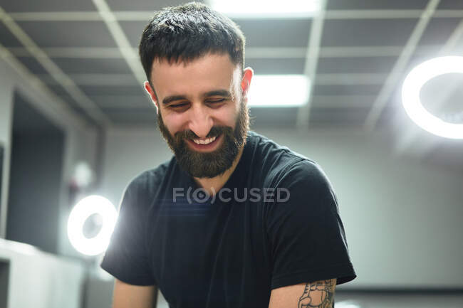 Happy bearded adult male tattooist in casual clothes sitting on stool near ring lamp and instruments while looking down in light tattoo salon — Stock Photo