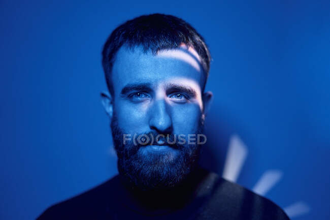 Selbstbewusster bärtiger erwachsener Mann in lässiger Kleidung blickt in dunklen Raum mit blauem Neonlicht und Schatten in die Kamera — Stockfoto