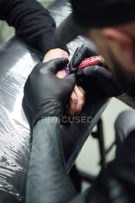 Concentrated male tattooist in gloves making tattoo on hand of client while using professional tattoo machine in modern tattoo studio — Stock Photo