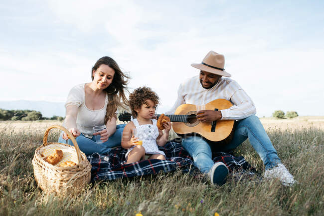 Delighted multiracial family enjoying picnic together while eating snacks and playing guitar in nature — Stock Photo