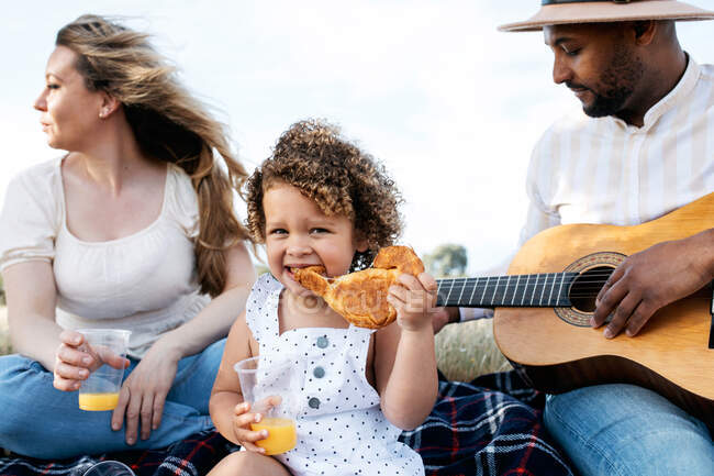 Encantada familia multirracial disfrutando de picnic juntos mientras comen bocadillos y tocan la guitarra en la naturaleza - foto de stock