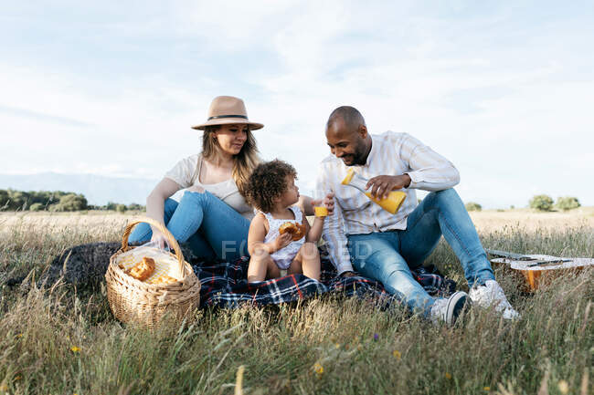 Zufriedenes multiethnisches Paar mit kleiner Tochter beim Picknick auf dem Feld, während es auf Plaid sitzt und Saft trinkt — Stockfoto