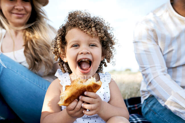 Fröhliches kleines Mädchen, das Gebäck isst und in die Kamera blickt, sitzt mit einer multiethnischen Familie und genießt ein gemeinsames Picknick in der Natur — Stockfoto