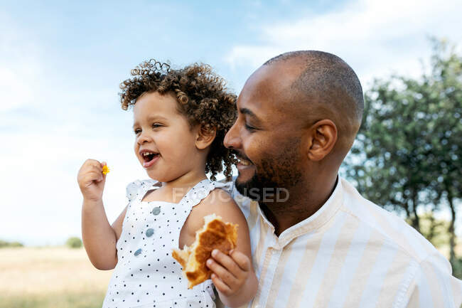 Позитивний батько афроамериканців, який обіймає чарівну дочку, споживаючи смачну булочку на літньому полі. — стокове фото