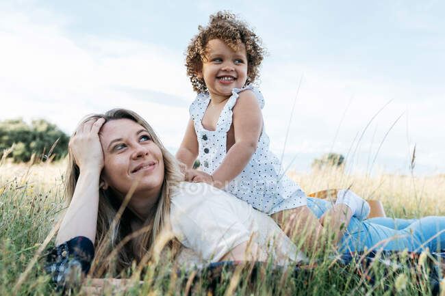 Side view of cheerful mother and curly haired little girl lying together on blanket in meadow and enjoying summer day — Stock Photo