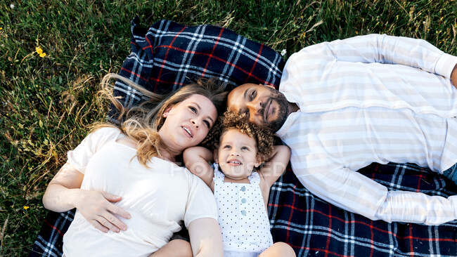 Зверху багатоетнічна сім'я з милою дитиною лежить разом на ковдрі на зеленій траві і мріє — стокове фото