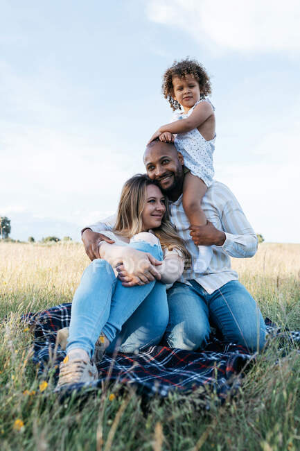 Vue latérale du couple multiracial joyeux avec adorable petite fille jouant dans la prairie en été — Photo de stock
