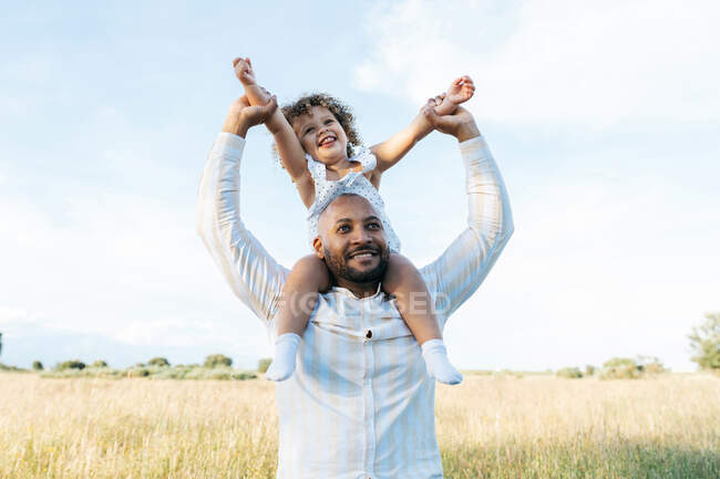 Веселий афроамериканець з милою донькою на плечах бавиться в полі влітку і веселиться, озираючись — стокове фото