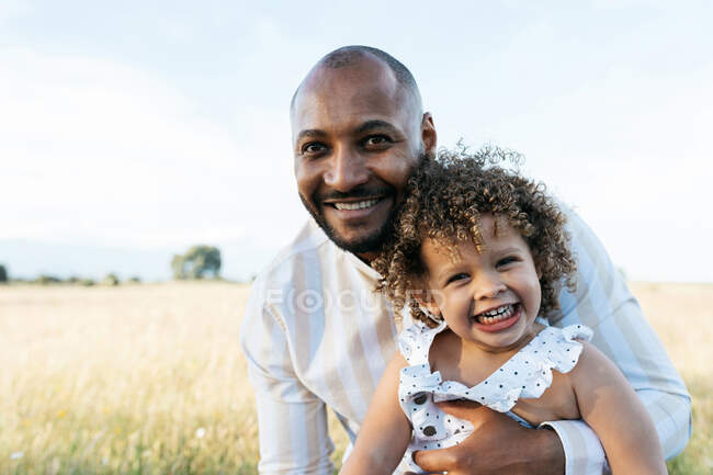 Африканський батько, який тримає симпатичну кучеряву афро-американську дочку на фоні сонячного неба в природі, дивлячись на камеру — стокове фото