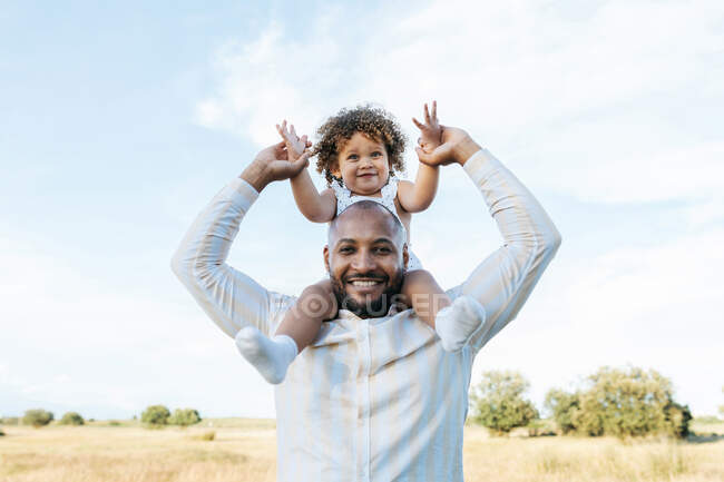 Cheerful African American father with cute little daughter on shoulders playing in field in summer and having fun looking at camera — Stock Photo