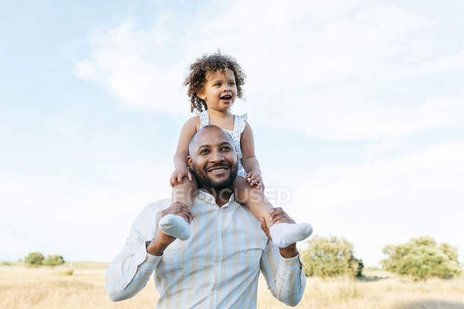 Веселый афроамериканец отец с милой маленькой дочкой на плечах играет в поле летом и весело смотрит в сторону — стоковое фото