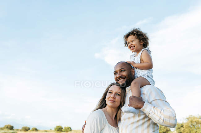 Niño sentado en los hombros del padre de pie en el campo con la madre en el día de verano - foto de stock