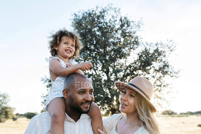 Child sitting on shoulders of father standing in field with mother on summer day — Stock Photo