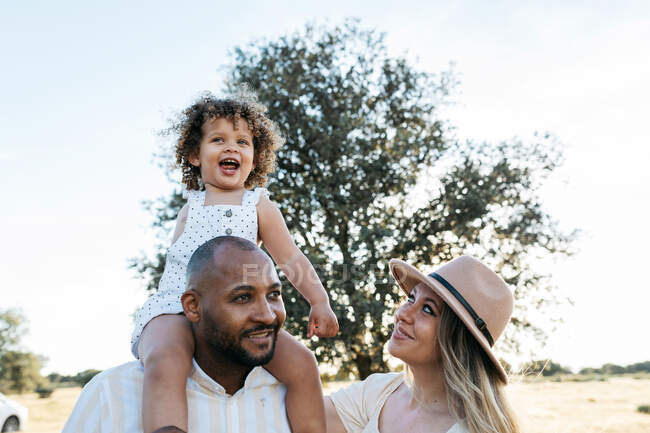 Child sitting on shoulders of father standing in field with mother on summer day — Stock Photo