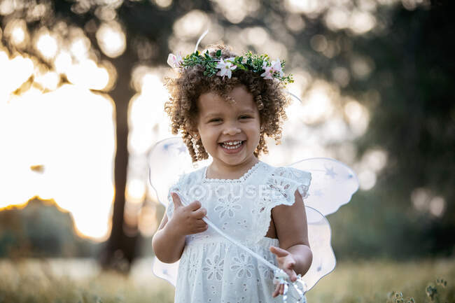 Entzücktes kleines Mädchen in Feenkostüm und Blumenkranz steht im Park und blickt in die Kamera — Stockfoto