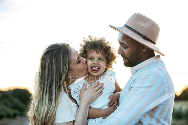 Delighted multiethnic family hugging cute little daughter while standing against sunset in field in summer — Stock Photo