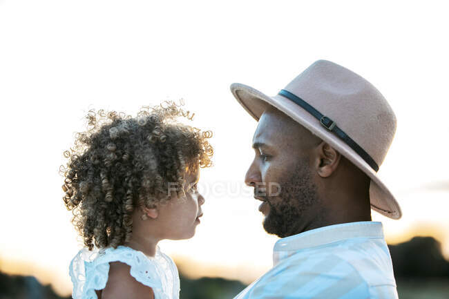 Vue latérale du père afro-américain tenant mignonne fille afro-américaine aux cheveux bouclés sur fond de ciel couchant dans la nature en se regardant — Photo de stock
