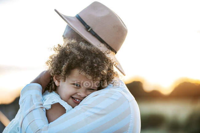 Vista lateral de un padre irreconocible abrazando a una linda hija afroamericana de pelo rizado en el fondo del cielo al atardecer en la naturaleza - foto de stock