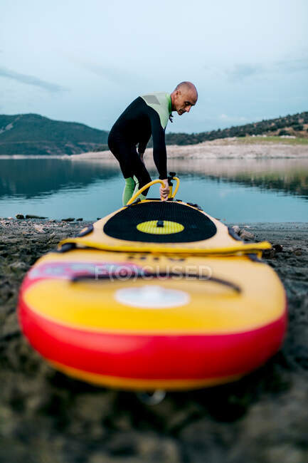 Männlicher Surfer im Neoprenanzug pumpt SUP-Board, während er am Strand steht und sich auf das Surfen vorbereitet — Stockfoto