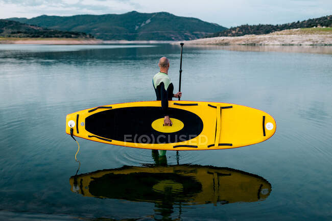 Side view of unrecognizable male surfer in wetsuit standing with yellow SUP board and paddle in sea water — Stock Photo