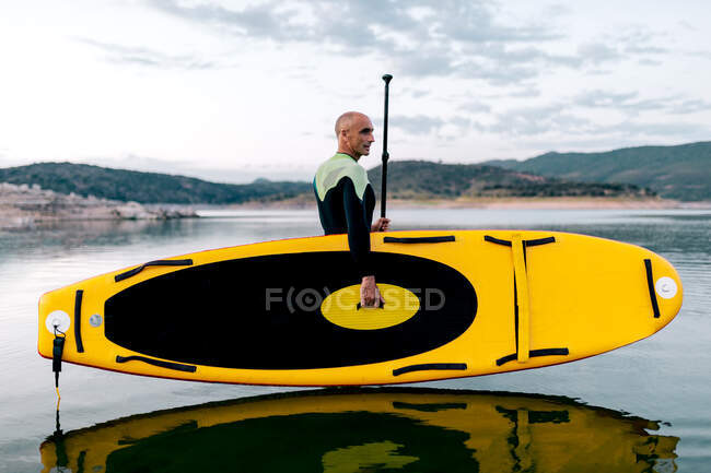 Vue latérale du surfeur masculin en combinaison debout avec SUP board jaune et pagaie dans l'eau de mer — Photo de stock