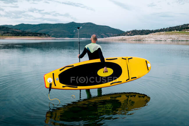 Вид сбоку на неузнаваемого серфера в гидрокостюме, стоящего с желтой доской SUP и веслом в морской воде — стоковое фото