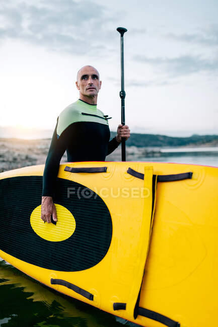 Vista lateral del surfista masculino en traje de neopreno de pie con tabla SUP amarilla y paleta en agua de mar - foto de stock