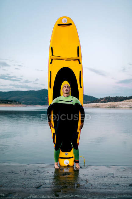 Calm male surfer in wetsuit standing eyes closed with yellow SUP board on beach near sea — Stock Photo