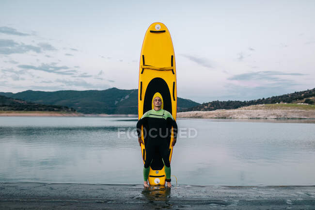 Calma surfista masculino en traje de neopreno de pie con los ojos cerrados con tabla SUP amarillo en la playa cerca del mar - foto de stock