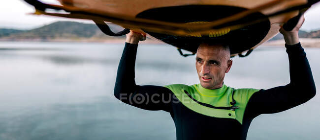 Mature male surfer standing with paddleboard above head near sea in evening and looking away — Stock Photo