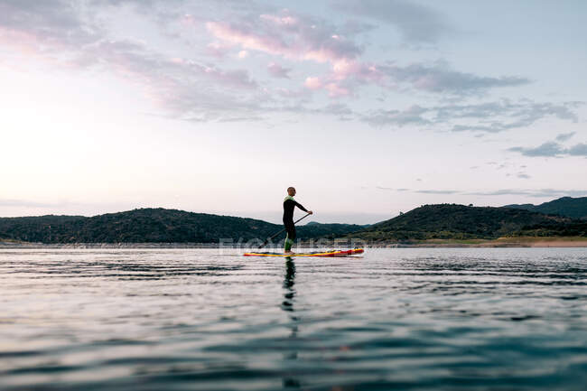 Seitenansicht eines anonymen männlichen Surfers, der bei Sonnenuntergang im Sommer auf einem SUP-Board auf ruhiger See schwimmt — Stockfoto