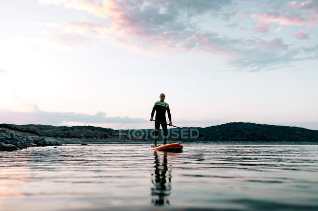 Von unten fokussierter männlicher Surfer, der bei Sonnenuntergang im Sommer auf dem SUP-Board auf ruhiger See schwimmt — Stockfoto