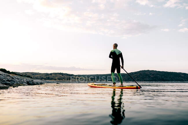 Back view of anonymous male surfer floating on SUP board on calm sea at sunset in summer — Stock Photo