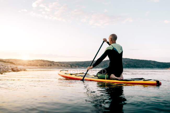 Full body side view of adult male in wetsuit kneeling on paddle board on calm lake water surface — Stock Photo