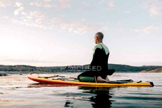 Vista lateral del tranquilo surfista masculino sentado en Thunderbolt posando en el paddleboard y mediando mientras practica yoga en el mar por la noche - foto de stock
