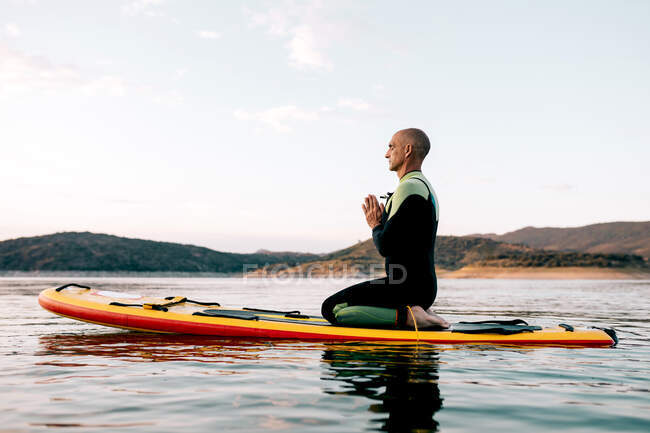 Seitenansicht des ruhigen männlichen Surfers, der in Thunderbolt-Pose mit Namaste Händen auf dem Paddleboard sitzt und vermittelt, während er abends Yoga im Meer praktiziert — Stockfoto
