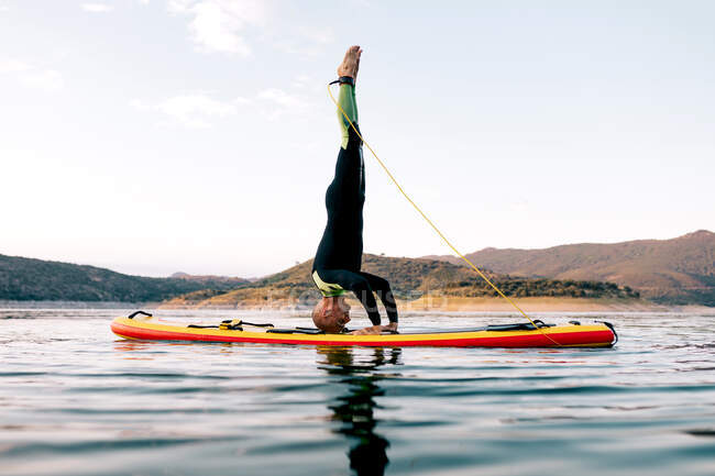 Vista laterale del surfista di sesso maschile praticare yoga in posa poggiatesta sulla SUP bordo in mare al tramonto — Foto stock