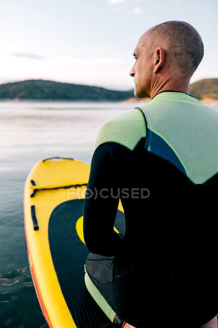 Rückenansicht eines erwachsenen Männchens im Neoprenanzug, das auf einem Paddelbrett auf der ruhigen Wasseroberfläche des Sees kniet — Stockfoto
